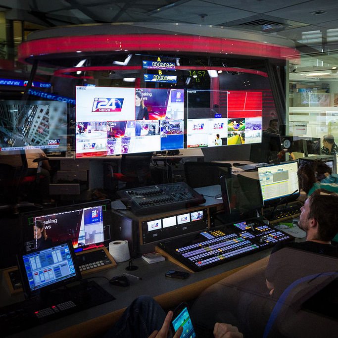 L'image montre une salle de contrôle de télévision ou de production, probablement un centre de diffusion. On y voit plusieurs écrans affichant des images en direct, des graphiques et des informations. Des personnes sont assises devant des consoles de contrôle, concentrées sur leur travail, et semblent superviser la diffusion. L'éclairage est tamisé, et on peut apercevoir une ambiance technologique avec des équipements modernes. Les murs en verre permettent d'apercevoir à l'extérieur, donnant une sensation d'ouverture.