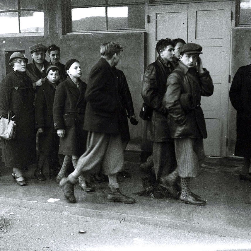 L'image montre un groupe de personnes, majoritairement des femmes et des enfants, marchant en direction d'un bâtiment. Ils portent des vêtements sombres et d'hiver. Leurs expressions semblent sérieuses et préoccupées. À l'entrée du bâtiment, une personne se tient à l'écart, observant le groupe avec une expression indéchiffrable. L'atmosphère de l'image est pesante et évoque un sentiment de tristesse ou d'incertitude. La scène semble avoir lieu dans un cadre urbain ou un lieu de rassemblement, avec des fenêtres en arrière-plan.