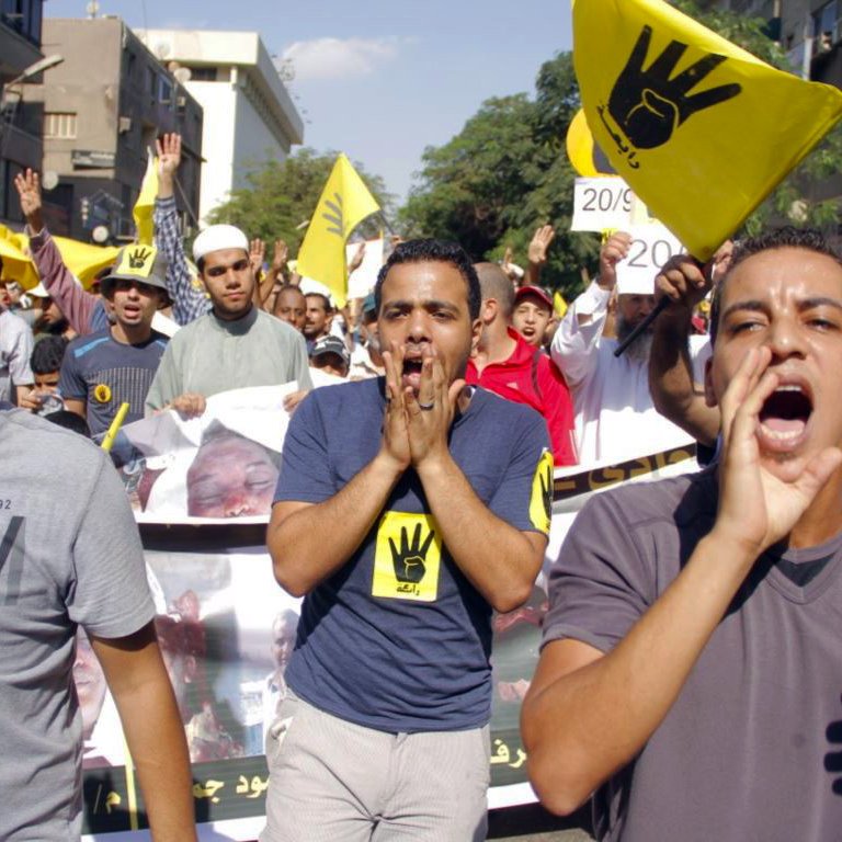 L'image montre une manifestation animée, avec des participants portant des t-shirts colorés. Ils expriment des slogans, certains scandent en levant les bras, tandis que d'autres tiennent des pancartes, notamment avec un symbole en forme de main. L'ambiance semble être pleine d'énergie et de détermination, reflétant une forte mobilisation autour d'une cause. Les bâtiments en arrière-plan suggèrent un milieu urbain.