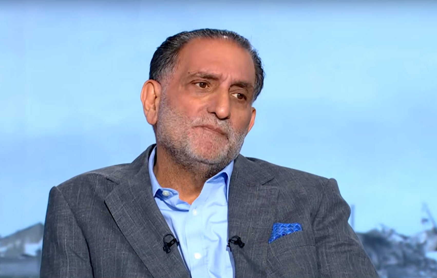 A middle-aged man in a suit with a serious expression, seated against a blurred background.