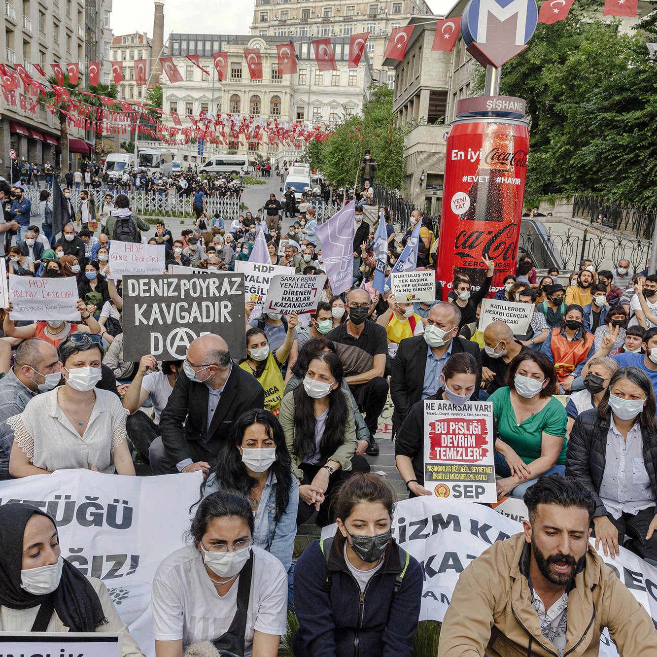 L'image montre une manifestation en plein air avec de nombreuses personnes rassemblées dans une rue. Les manifestants portent des masques et brandissent des pancartes avec des slogans. L'atmosphère semble mobilisée, et il y a beaucoup de drapeaux en arrière-plan. Les participants sont assis pour exprimer leur message, signalant une volonté de protester pacifiquement.