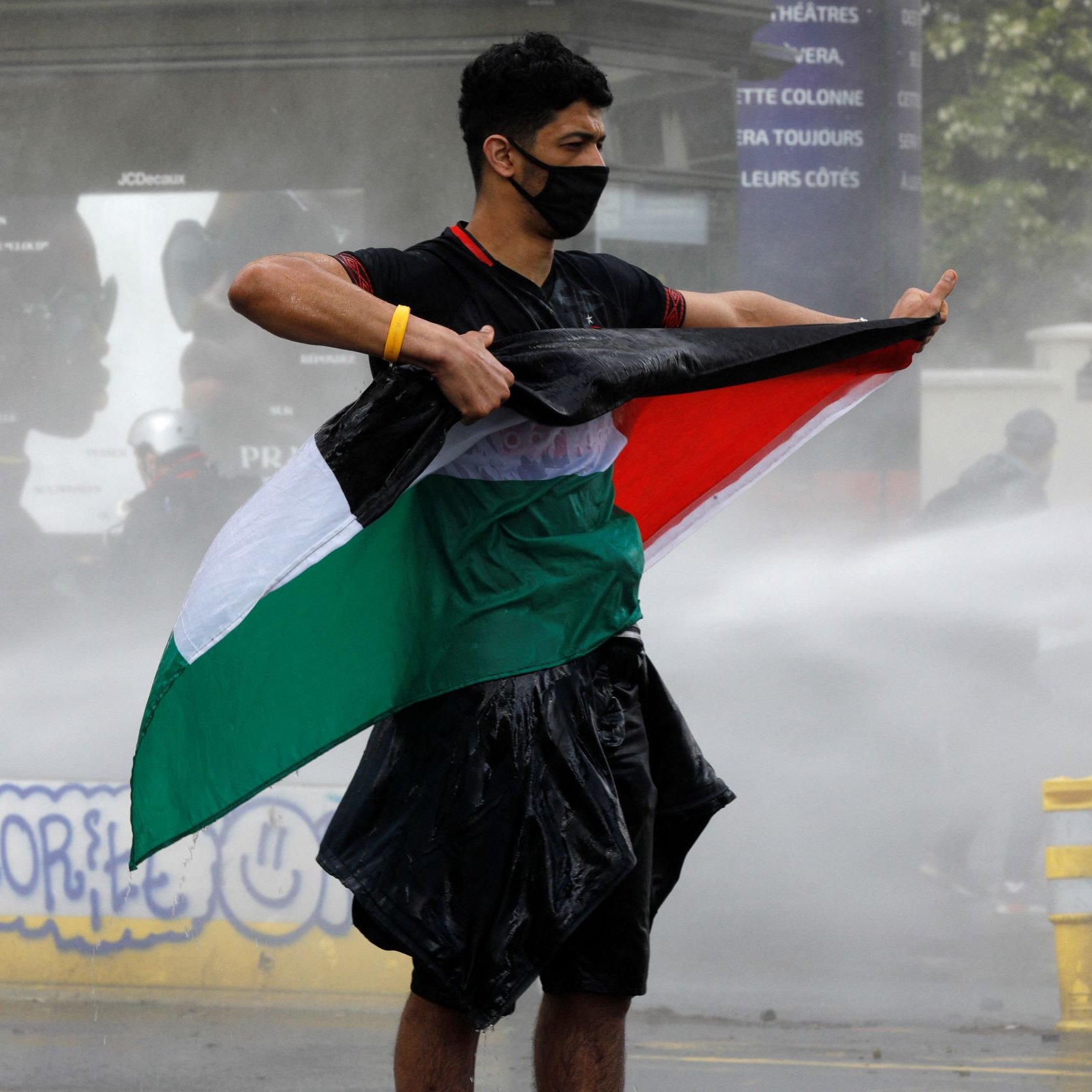 Sur l'image, on voit un jeune homme debout, tenant un drapeau. Il porte un masque noir et semble faire face à une situation de tension, peut-être une manifestation. Le drapeau qu'il brandit a des couleurs verde, blanc et rouge, tandis que de l'eau jaillit autour de lui, suggérant l'utilisation de canons à eau. L'environnement semble être urbain, avec des signes de protestation en arrière-plan.