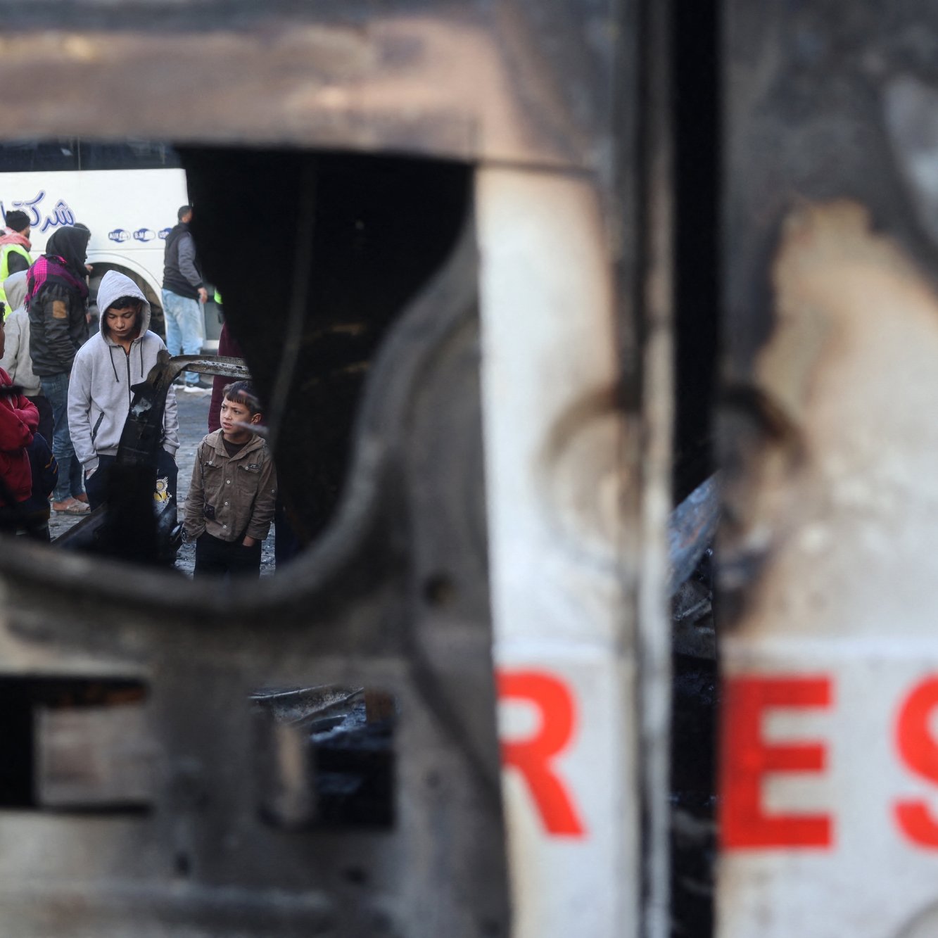 L'immagine mostra una scena cupa e difficile, probabilmente in un contesto di conflitto o crisi. Si vede un gruppo di persone che si trovano in un'area devastata, ricevendo assistenza o cercando di mettersi in salvo. Parte della scena è incorniciata da un oggetto danneggiato, che contribuisce a esprimere la tensione e la difficoltà della situazione. Le persone nel gruppo appaiono preoccupate e potrebbero essere in attesa di aiuto o di informazioni.