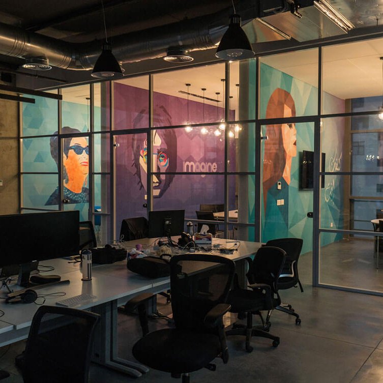 L'image montre un espace de bureau moderne avec de grandes fenêtres offrant une vue sur l'extérieur. Les murs sont décorés de peintures murales colorées représentant des visages stylisés. Le mobilier est minimaliste et fonctionnel, avec des tables de travail et des chaises ergonomiques. L'éclairage est tamisé, créant une ambiance chaleureuse malgré le design contemporain. On peut également voir des ordinateurs sur les bureaux, indiquant que l'espace est utilisé pour le travail.