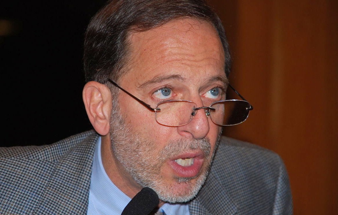 Je ne sais pas qui est cette personne. Cependant, je peux dire que l'image semble montrer un homme portant des lunettes, avec une barbe grisonnante et des cheveux également grisonnants. Son expression semble sérieuse et il est peut-être en train de parler ou de faire une présentation. Son veston est en tissu à motifs.