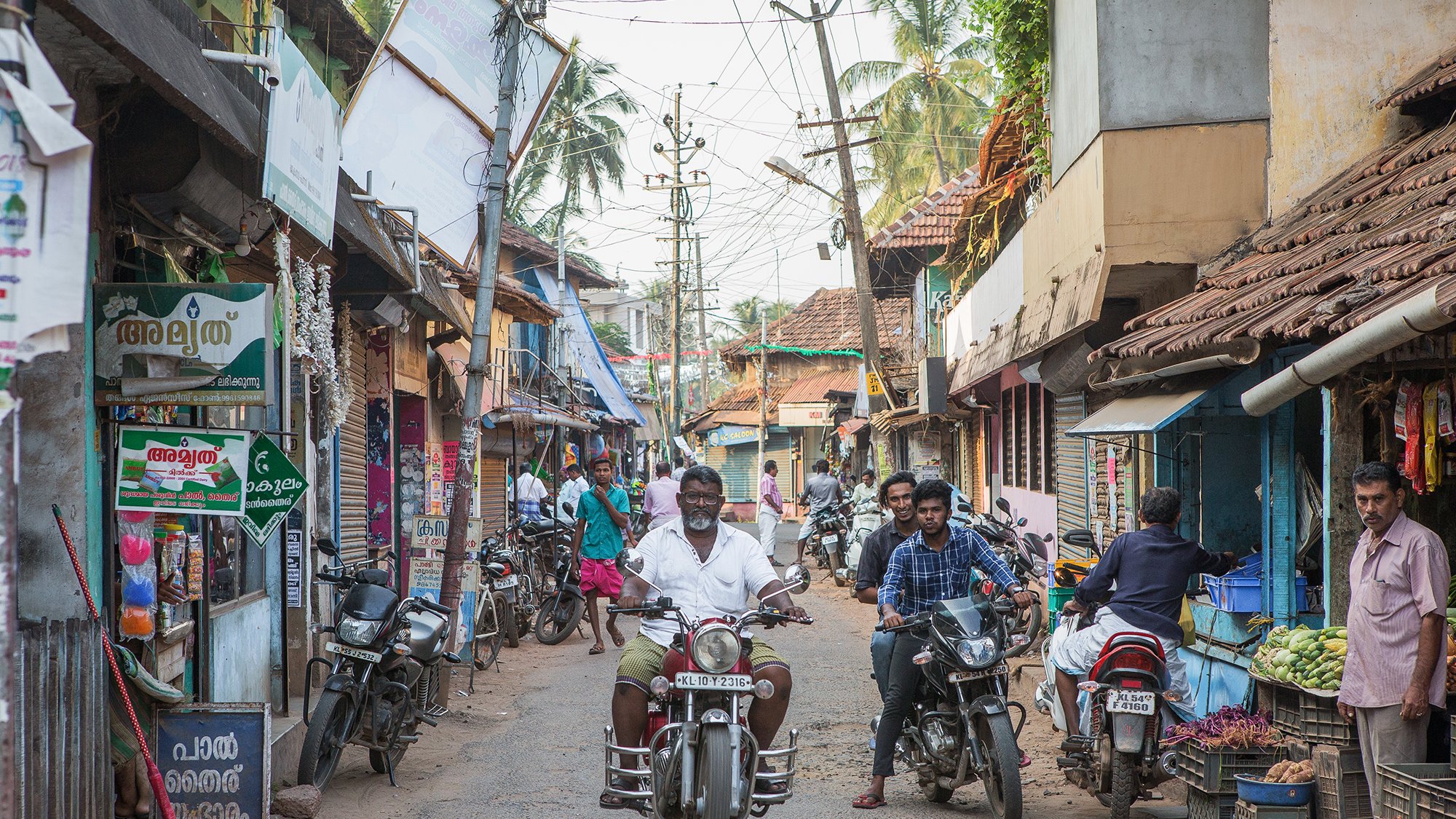 تظهر الصورة مشهداً من شارع ضيق في منطقة ريفية أو شبه حضرية. يتواجد في الصورة عدد من الأشخاص يسيرون ويتحدثون، بينما يقود رجل دراجة نارية في منتصف الشارع. المباني على الجانبين بسيطة ومكونة من طوابق قليلة، مع أسطح مغطاة بالبلاط. يمكن رؤية لافتات ملونة على المحلات التجارية التي تبيع مختلف السلع، وهناك أشجار تتواجد في الخلفية مما يعطي انطباعاً بحياة محلية نابضة. الجو يبدو هادئاً ومعتاداً على النشاط اليومي للسكان.