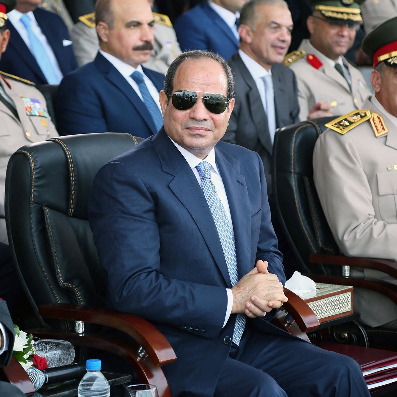 L'image montre un groupe de personnes lors d'un événement officiel. Au premier plan, un homme portant des lunettes de soleil et habillé d'un costume sombre est assis sur une chaise, semblant attentif. Il y a d'autres hommes en uniforme militaire et en tenue formelle derrière lui, qui semblent également être présents pour l'événement. L'ambiance est solennelle et il semble s'agir d'une cérémonie ou d'une célébration officielle.