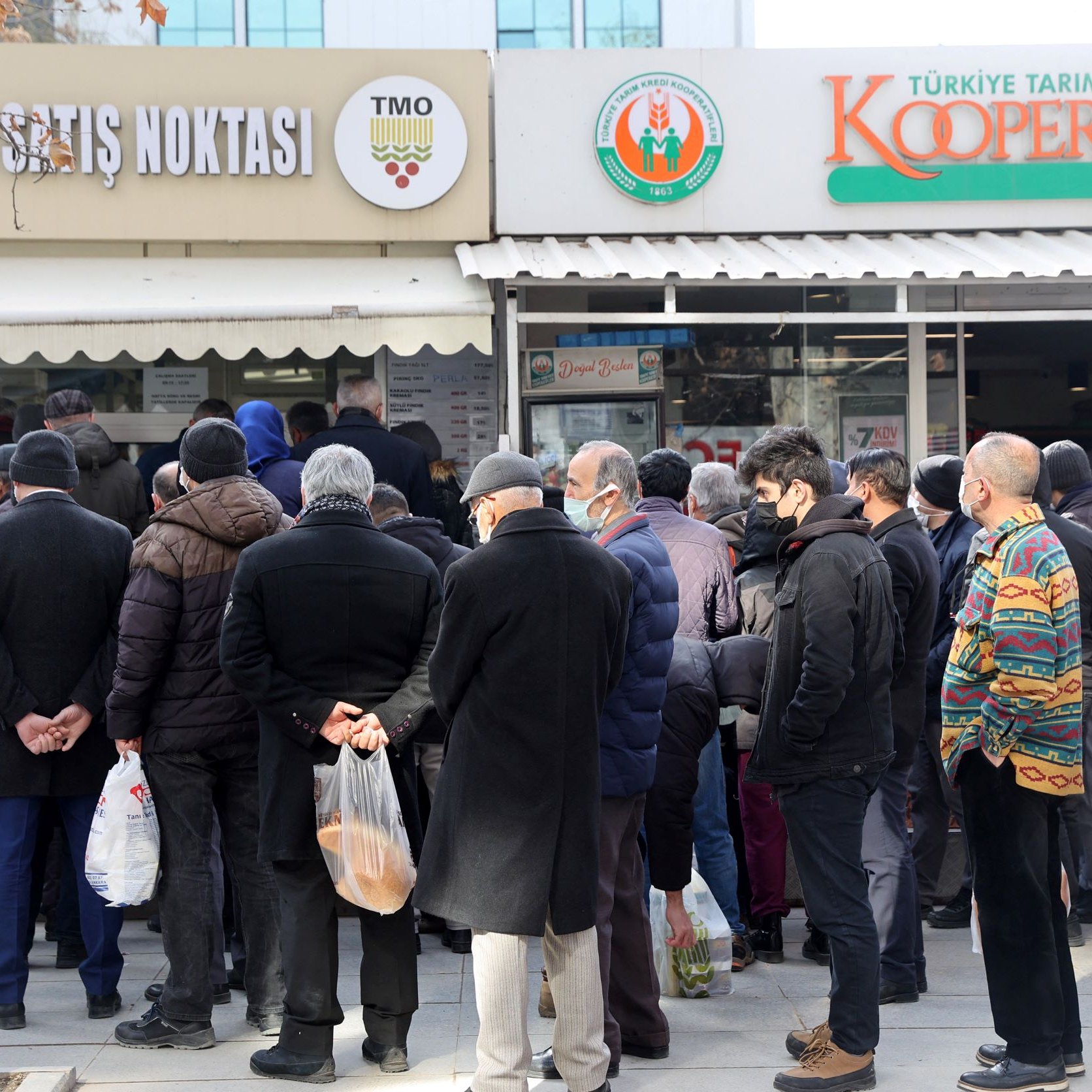 L'image montre une foule de personnes faisant la queue devant un magasin, probablement un marché ou un point de vente. Les gens semblent attendre pour entrer ou acheter des produits. Ils portent des vêtements d'hiver, et certains tiennent des sacs. L'atmosphère semble calme, mais il y a une certaine impatience dans la file d'attente. Au-dessus de l'entrée du magasin, on peut voir des panneaux indiquant le nom et la fonction de l'établissement.