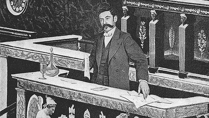 در این تصویر، مردی با لباس رسمی در حال ایستادن در مقابل یک میز بزرگ است. پس‌زمینه شامل طراحی‌های تزئینی و نقوش هنری می‌باشد. بر روی میز اشیایی مانند یک ظرف و مدارکی قرار دارد. در دو طرف میز، دو مجسمه فرشته وجود دارد که در حال نوشتن و نواختن ساز هستند. فضای کلی تصویر تاریخی و با وقار به نظر می‌رسد.