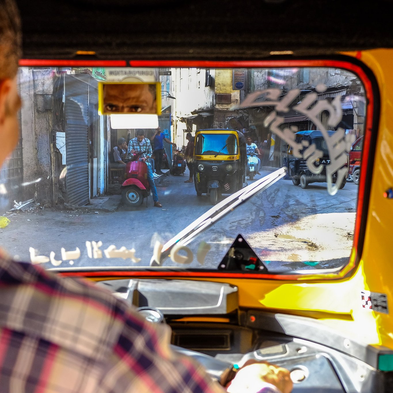 L'image montre la vue depuis l'intérieur d'un tuk-tuk, avec un conducteur visible en premier plan. On aperçoit des rues étroites et animées, avec d'autres véhicules, y compris un autre tuk-tuk. Des bâtiments anciens bordent la rue, et il y a des détails comme des affiches et des éléments de la vie urbaine qui ajoutent du caractère à l'environnement. La lumière naturelle éclaire la scène, créant une atmosphère vivante et dynamique.