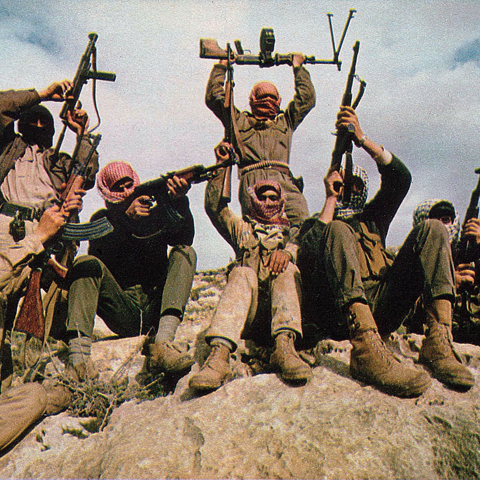 الصورة تظهر مجموعة من الأشخاص يرتدون ملابس عسكرية، وهم يتجهون نحو الكاميرا. بعضهم يحمل بنادق، بينما يقوم شخص في المنتصف برفع سلاح فوق رأسه. يظهر الأشخاص في وضعية متحمسة على صخرة كبيرة، وتظهر الخلفية سماء زرقاء مع بعض الغيوم. تعبّر الصورة عن روح الرفاقية والتصميم.