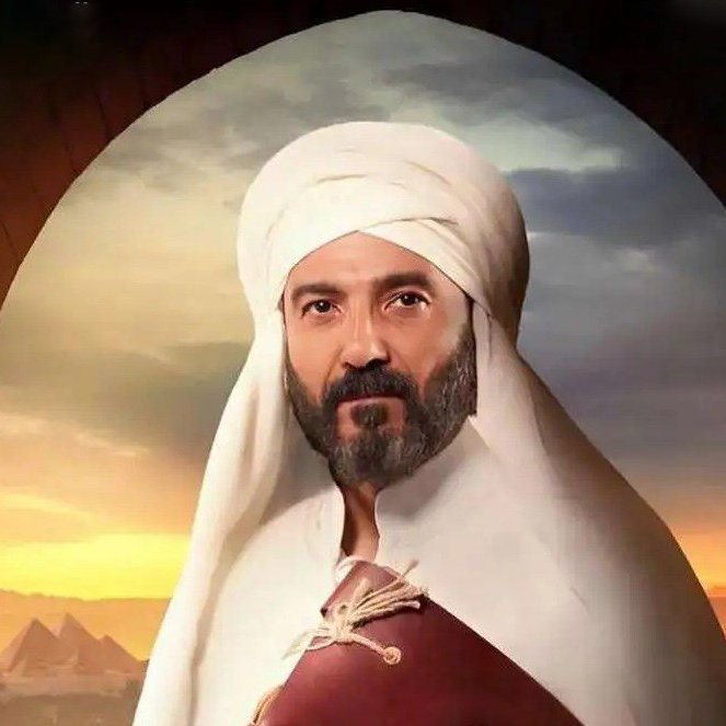 L'image représente un homme portant un turban et une tenue traditionnelle. Il a une barbe et une expression sérieuse. En arrière-plan, on aperçoit un paysage désertique avec des pyramides, suggérant une ambiance historique ou culturelle. La lumière semble douce, ajoutant une atmosphère dramatique à la scène.