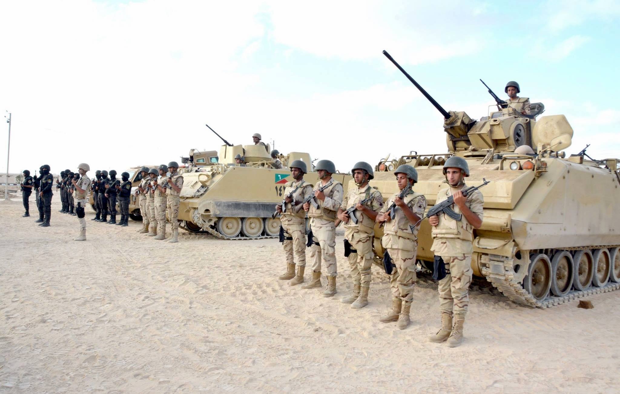 تظهر في الصورة مجموعة من الجنود العسكريين يقفون في صف منتظم، يرتدون زيًا عسكريًا ويحملون أسلحة. خلفهم توجد مدرعات عسكرية، وبعض الجنود يجلسون فوق هذه المدرعات. يبدو أن المشهد قد تم التقاطه في منطقة صحراوية أو رملية، مع سماء صافية في الخلفية. تعكس الصورة أجواءً عسكرية منظمّة وجادة.