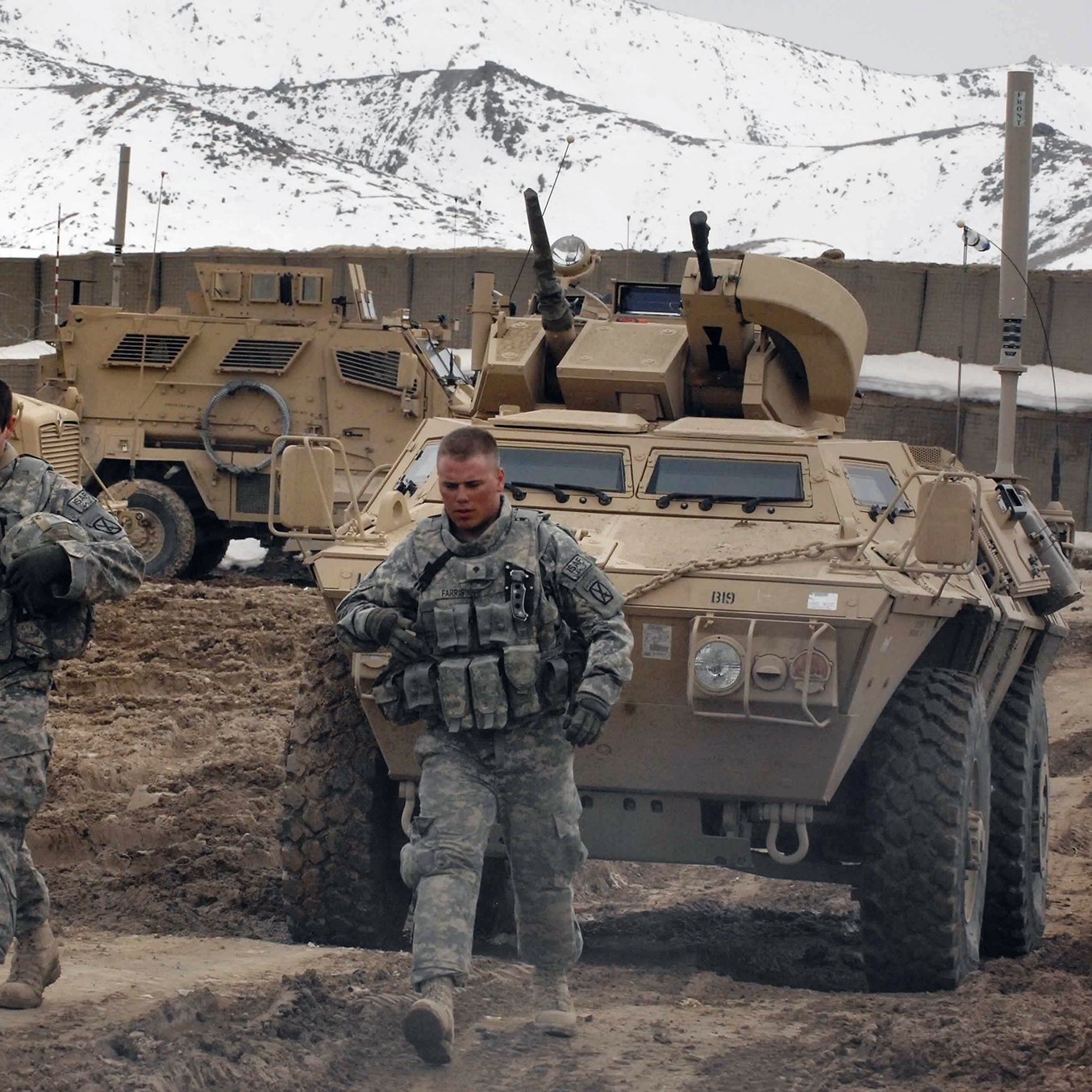 تظهر الصورة مشهدًا عسكريًا في منطقة جبلية. في الصورة، يسير جنديان في زي عسكري في المقدمة، مع التركيز على تفاصيل ملابسهم ومعداتهم. خلفهما، توجد مركبات مدرعة تُستخدم في العمليات العسكرية، محاطة بتضاريس جبلية تغطيها الثلوج. يبدو أن الطقس بارد، ويعكس المشهد البيئة الصعبة التي يعمل فيها الجنود.