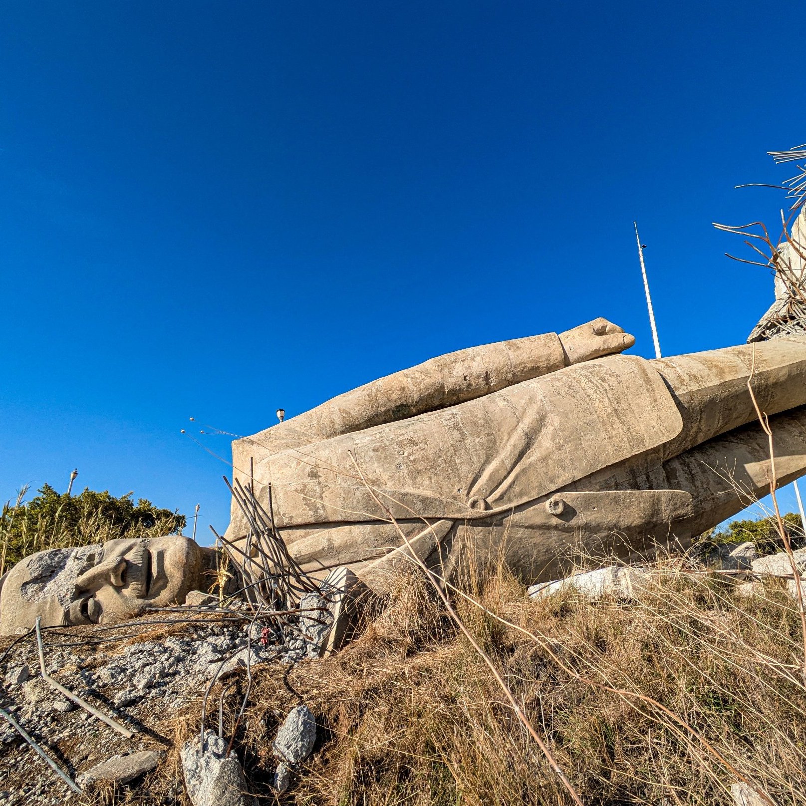 L'image montre une statue gigantesque de la main et du visage d'un personnage allongé sur le sol, entourée de verdure et sous un ciel bleu. La statue semble partiellement détruite ou en ruine.