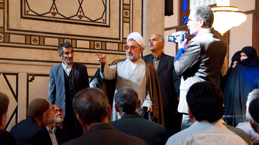 تظهر الصورة مجموعة من الأشخاص في مكان داخلي، حيث يركز المشهد على رجل يرتدي لباسًا تقليديًا يشير بيده ويتحدث إلى الحضور. يبدو هناك تفاعل قوي بين المتحدث والجمهور، الذين يظهرون جالسين أو واقفين في صفوف أمامه. في الخلفية، يمكن رؤية بعض الأشخاص الآخرين، وبعضهم يرتدي ملابس تقليدية. الأجواء تعكس طابعًا ثقافيًا أو دينيًا، مع تفاصيل معمارية تزين الجدران.