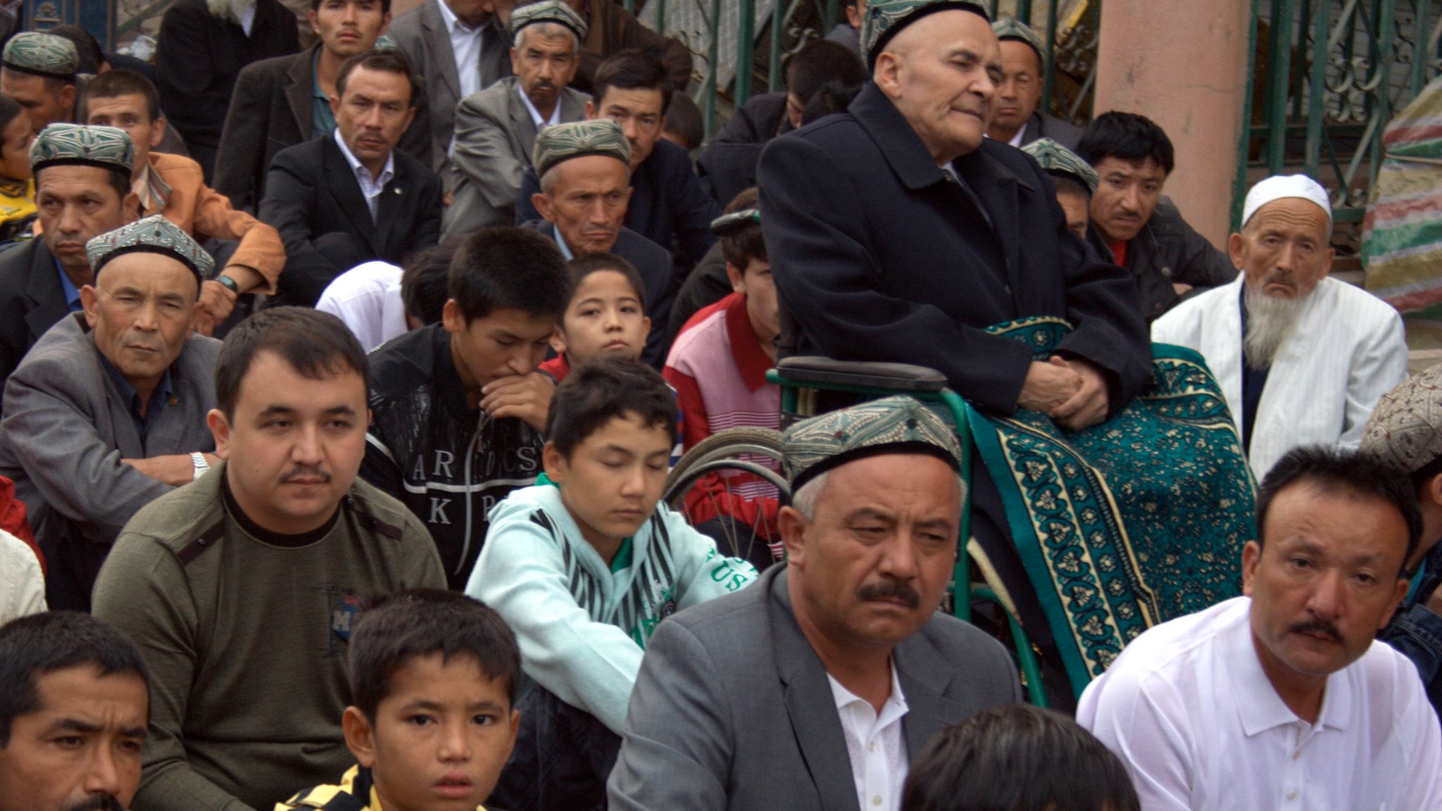 The image depicts a group of individuals seated together, primarily men, in a communal setting. They appear to be engaged in a gathering or possibly a religious ceremony, with some wearing traditional attire. In the foreground, there is an elderly man seated in a wheelchair, and others are attentively looking forward. The overall atmosphere reflects a sense of community and shared purpose. The individuals show a range of ages, from children to older men, indicating that the event likely holds significance for various generations.