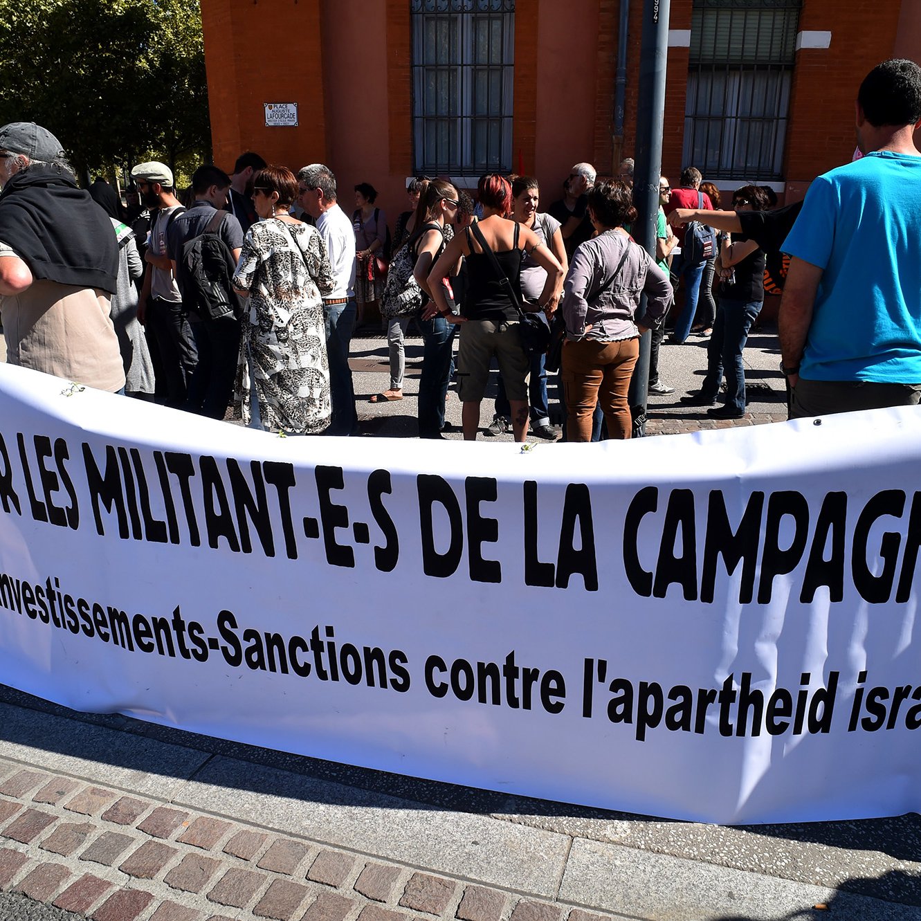 L'image montre un groupe de personnes rassemblées devant une bannière. La bannière, qui est en français, demande un soutien pour les militants de la campagne BDS, qui signifie Boycott, Désinvestissement et Sanctions contre l'apartheid israélien. Les manifestants semblent engagés et attentifs, participant à une action de protestation. Le décor urbain en arrière-plan indique que l'événement se déroule dans une ville, probablement en plein air.