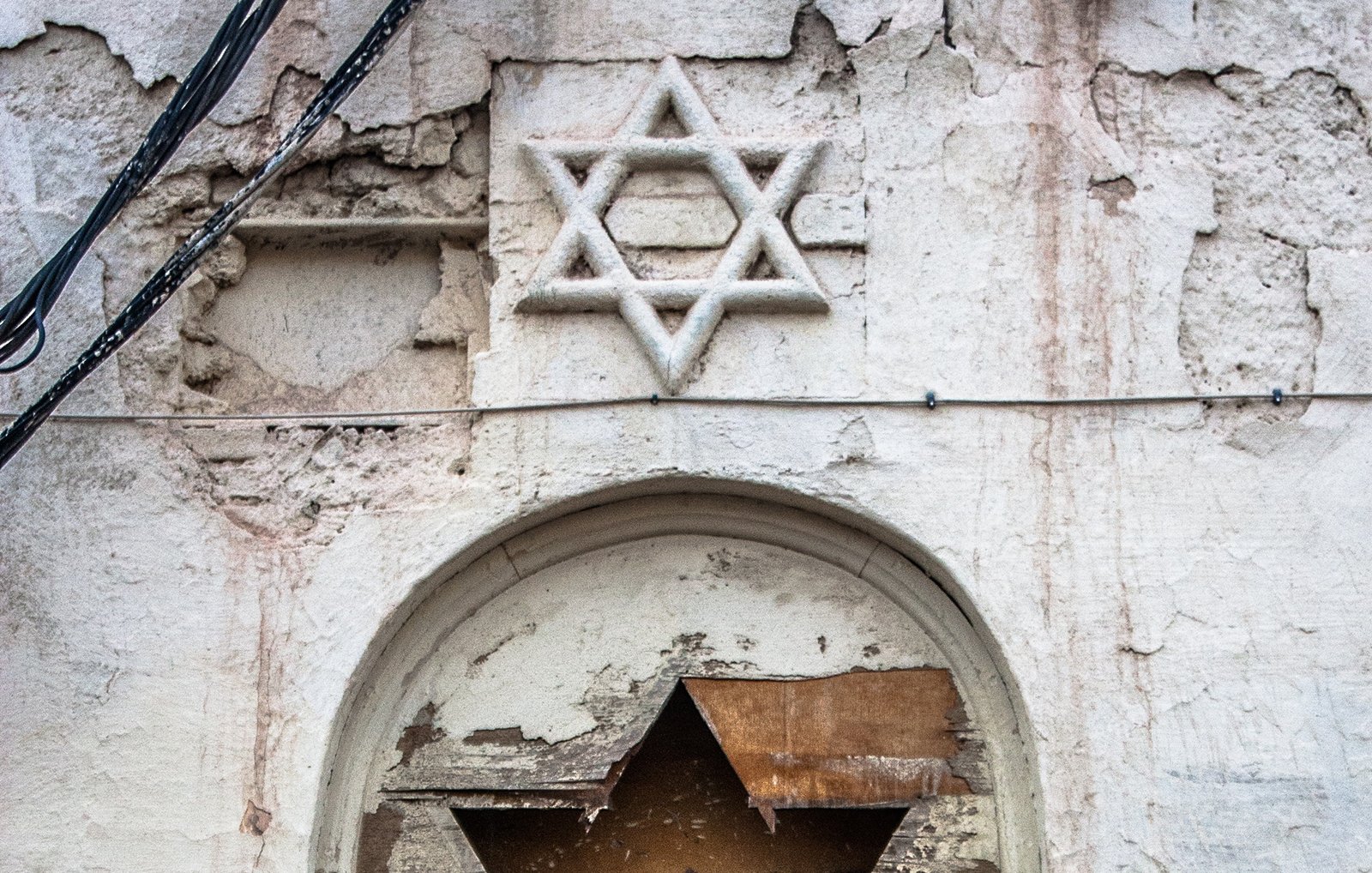 L'image montre un mur vieilli et abîmé, d'où une étoile de David est visible en haut, gravée ou ornée sur la surface. En dessous, une niche ou un cadre semble avoir été endommagé, avec des éléments en bois en mauvais état, suggérant un sentiment d'abandon. Un morceau de tissu bleu pend à l'intérieur de cette structure, ajoutant une touche de couleur à l'ensemble terni du mur. Les détails de la peinture écaillée et des fissures renforcent l'impression de délabrement et d'histoire.