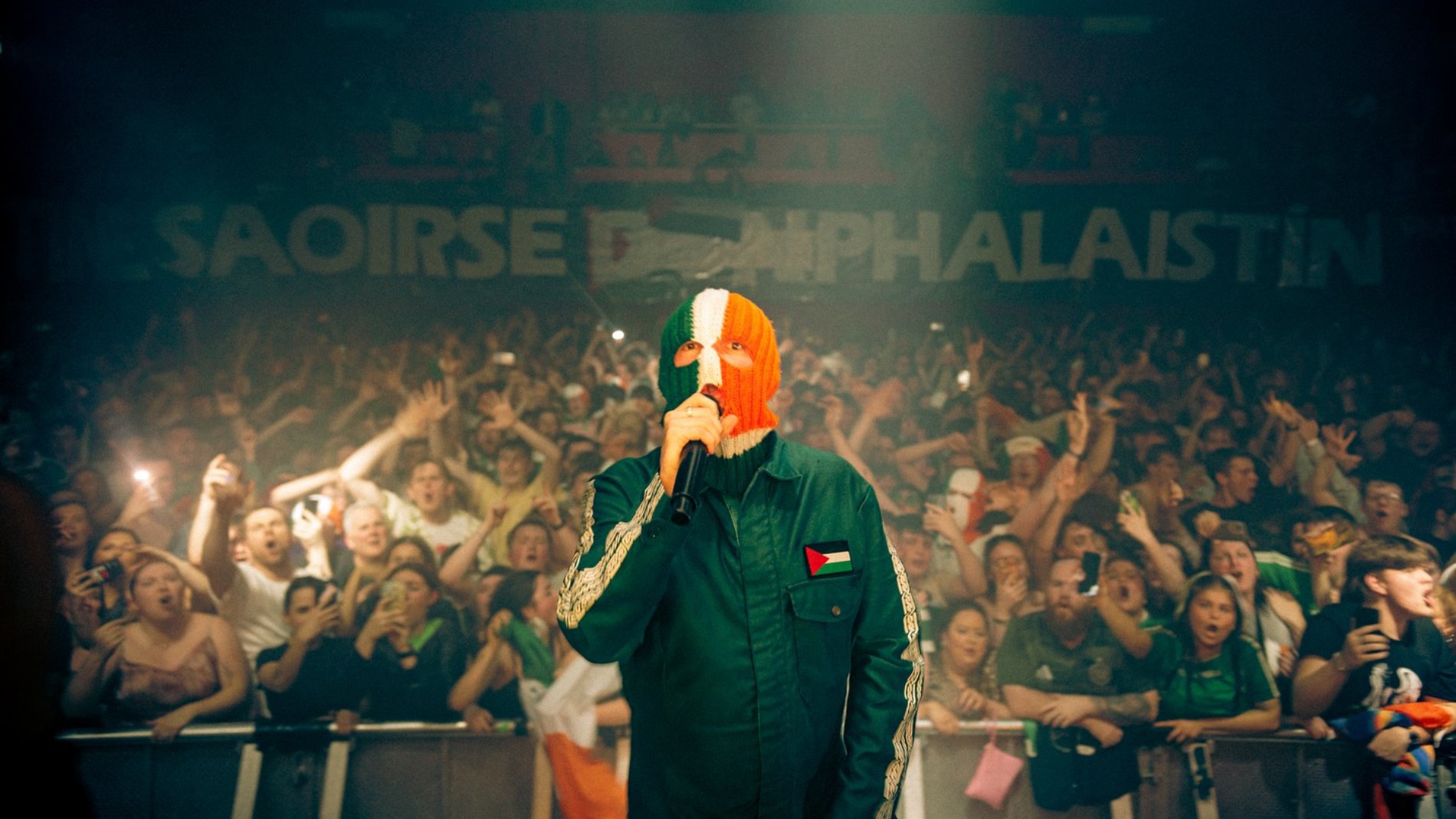 Un artiste en concert, masqué, devant une foule enthousiaste, avec des drapeaux irlandais et palestiniens.