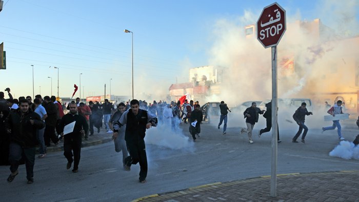 L'image montre une scène de manifestation. De nombreuses personnes courent dans une rue, entourées de fumée, probablement due à des gaz lacrymogènes. Au premier plan, on peut voir un panneau de signalisation "STOP". L'atmosphère semble tendue, et les manifestants semblent fuir une situation chaotique. Les participants portent des vêtements variés et certains tiennent des drapeaux.
