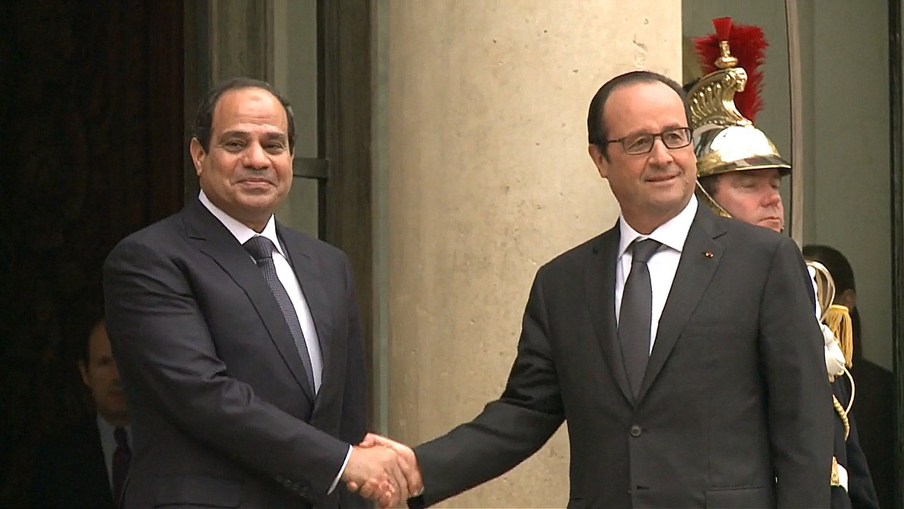 L'image montre deux hommes en costume, serrant la main l'un de l'autre. L'un est à gauche et semble avoir une expression amicale, tandis que l'autre se trouve à droite, affichant également un sourire. En arrière-plan, on aperçoit un soldat en uniforme. L'environnement semble être formel, probablement devant un bâtiment gouvernemental.