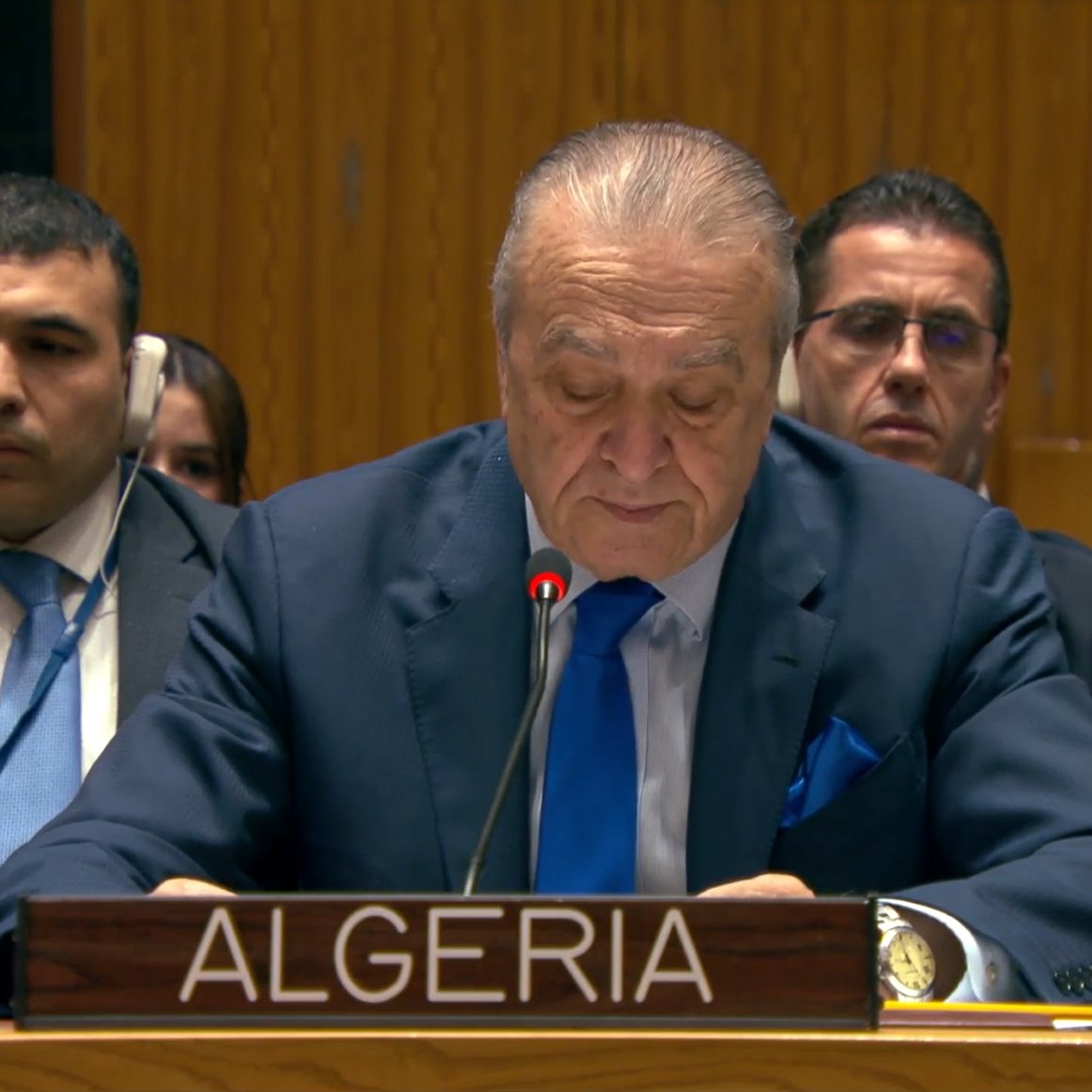 رجل يتحدث خلف منصة، يحمل لافتة الجزائر، مع أشخاص يجلسون خلفه في قاعة.