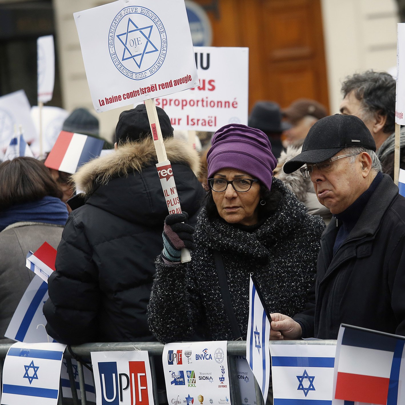 در این تصویر، گروهی از افراد در حال اعتراض هستند. آن‌ها پلاکاردها و تابلوهای مختلفی در دست دارند. برخی از افراد لباس‌های گرم به تن دارند و در میان آن‌ها یک زن با عینک دیده می‌شود. تابلوها شامل پیام‌هایی مرتبط با اسرائیل هستند و برخی از افراد به سمت دوربین نگاه می‌کنند. فضای تجمع در یک مکان عمومی به نظر می‌رسد.