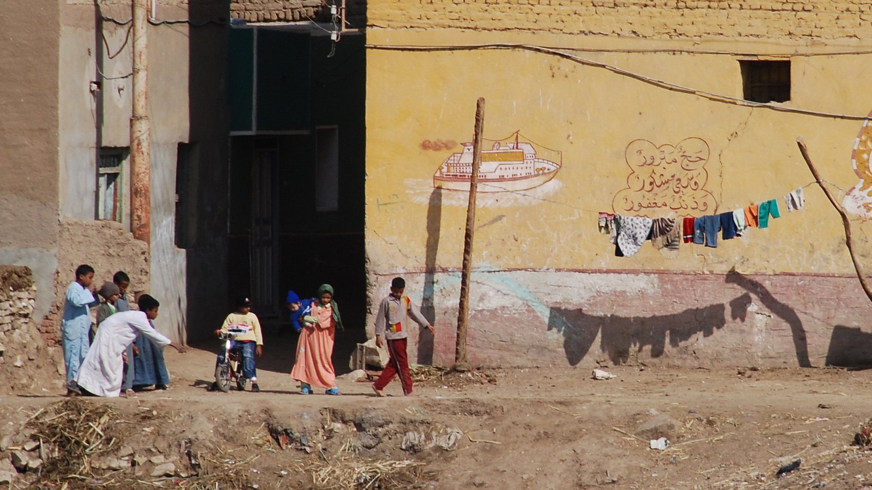 L'image montre une scène de rue dans un environnement rural ou semi-urbain. On peut y voir plusieurs personnes, dont des enfants, qui se déplacent. À l'arrière-plan, il y a des bâtiments avec des murs peints, incluant des motifs ou des inscriptions. Des vêtements sont suspendus à une corde, presque comme un étendage. Le sol est poussiéreux, et l'ensemble de la scène dégage une atmosphère de vie quotidienne dans un cadre modeste.
