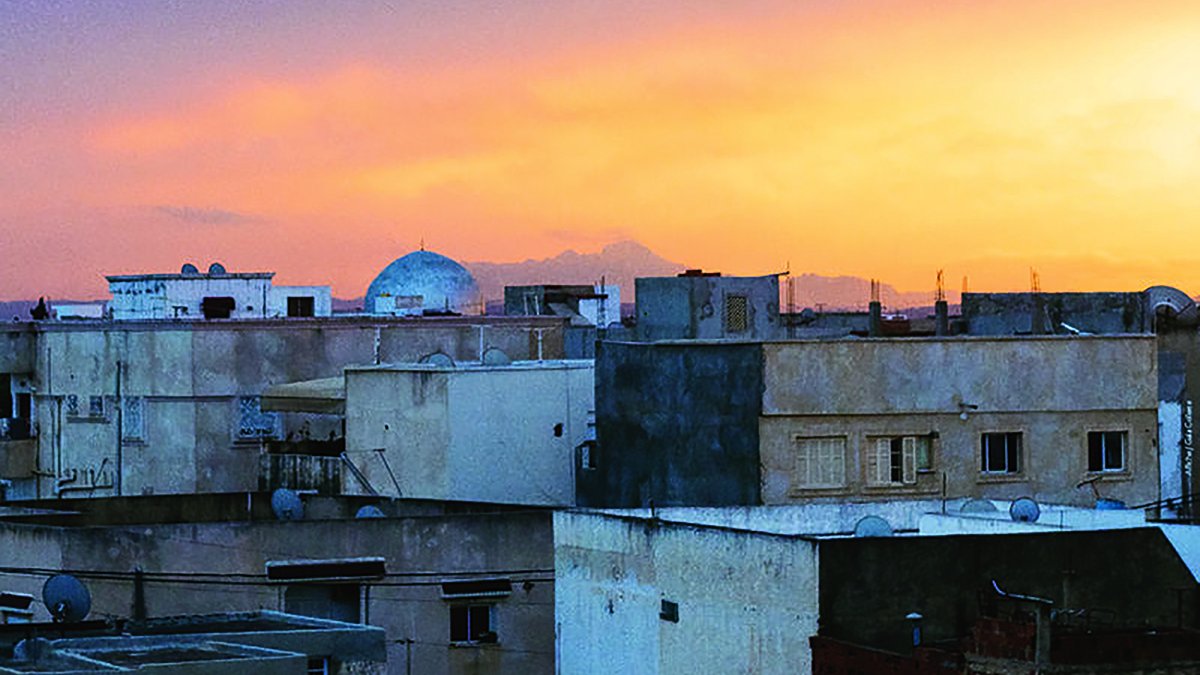 L'image montre un paysage urbain au coucher du soleil. On y voit plusieurs bâtiments aux façades de couleur beige et grise, avec des toits plats. À l'horizon, une belle teinte orange et rose illumine le ciel, créant une ambiance chaleureuse. Un dôme se distingue parmi les structures, suggérant la présence d'une mosquée ou d'un autre bâtiment significatif. Les lignes des constructions et le ciel coloré contrastent magnifiquement, donnant une impression de tranquillité.