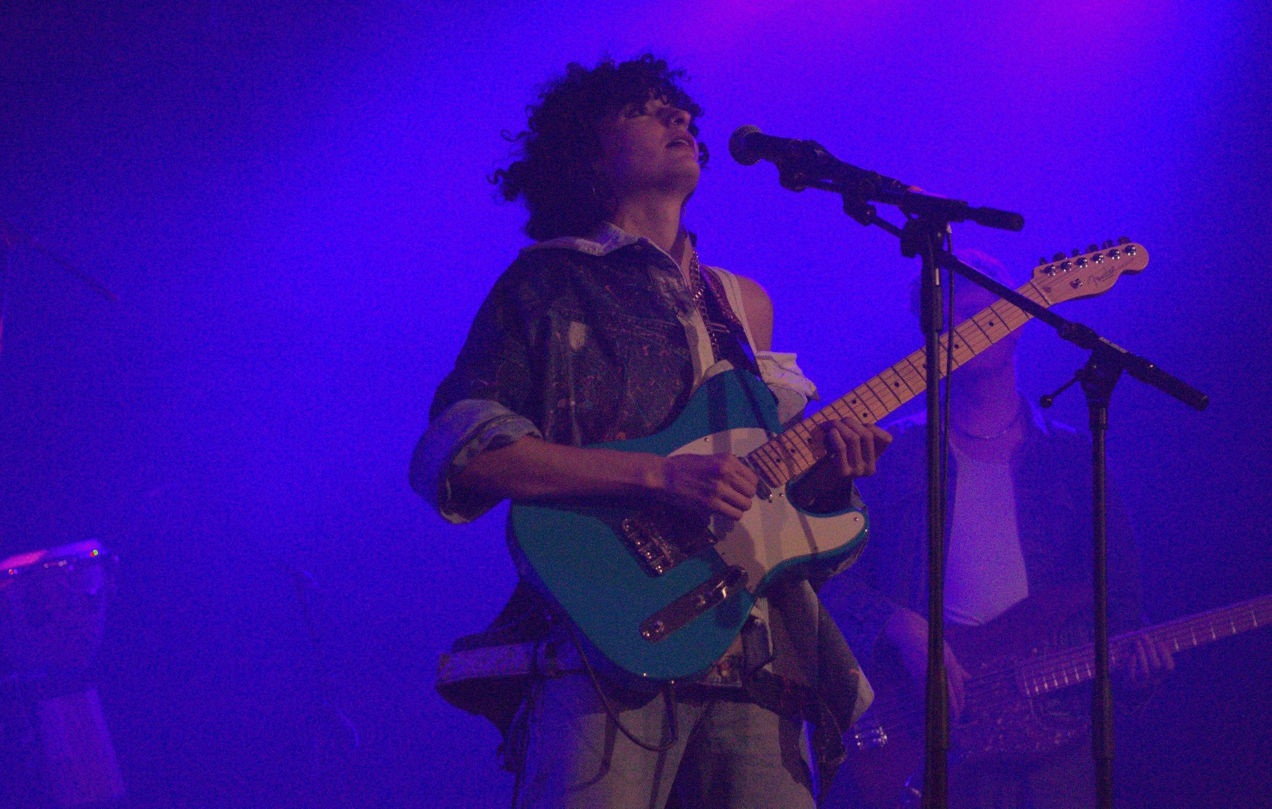 L'immagine mostra un musicista sul palco, intento a suonare una chitarra elettrica di colore azzurro. Ha i capelli ricci e indossa una camicia ampia con un look casual. Dietro di lui si intravedono altri musicisti e luci bluastre che creano un'atmosfera suggestiva. L'artista sembrano concentrato e coinvolto nella sua performance.