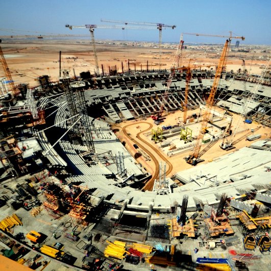 L'image montre un immense chantier de construction d'un stade. On peut voir une grande arène en forme de cercle, avec des gradins en cours d'assemblage. De nombreuses grues sont présentes, surélevant des matériaux de construction, et divers engins lourds sont dispersés sur le site. À l'horizon, on aperçoit des bâtiments et un ciel dégagé, indiquant une propreté et un bon déroulement des travaux. L'ensemble de la scène illustre l'activité dynamique et l'envergure d'un projet architectural ambitieux.