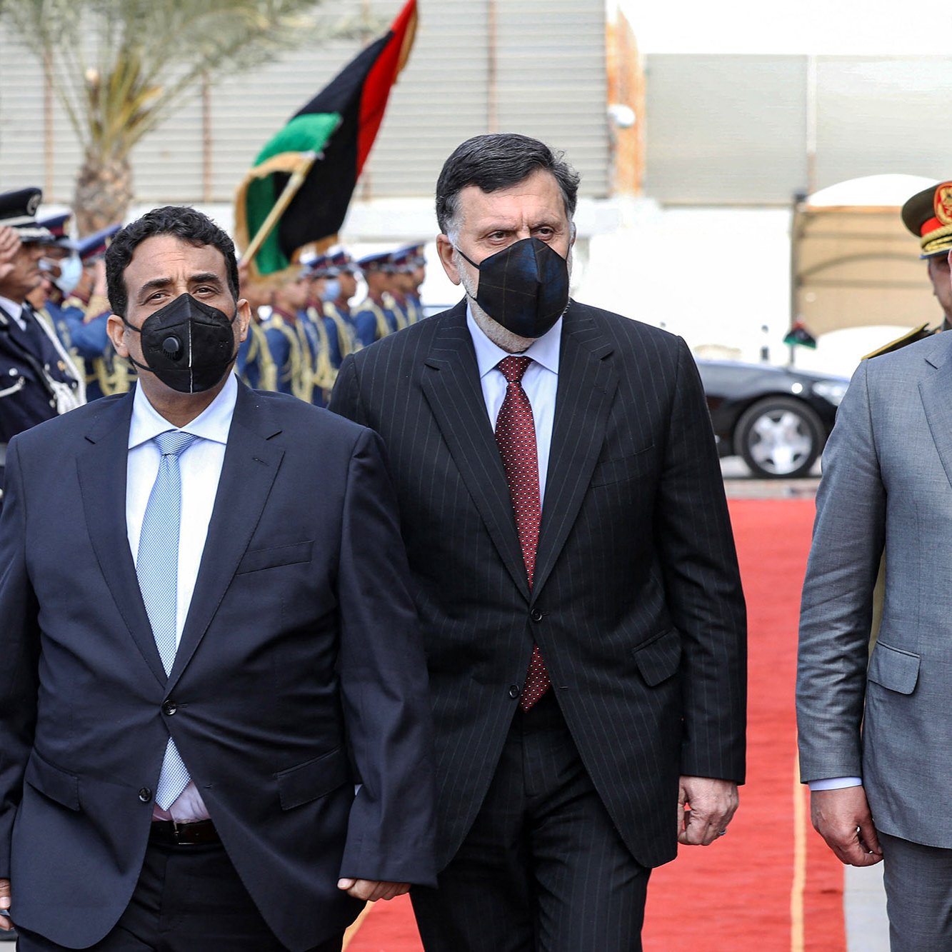 L'image montre un groupe de trois hommes marchant sur un tapis rouge, apparemment lors d'un événement officiel. Ils portent tous des costumes, et deux d'entre eux portent des masques faciaux noirs. En arrière-plan, on peut voir une garde d'honneur en uniforme, ainsi que des drapeaux. L'atmosphère semble solennelle et accueillante, suggérant une réception ou une cérémonie diplomatique.