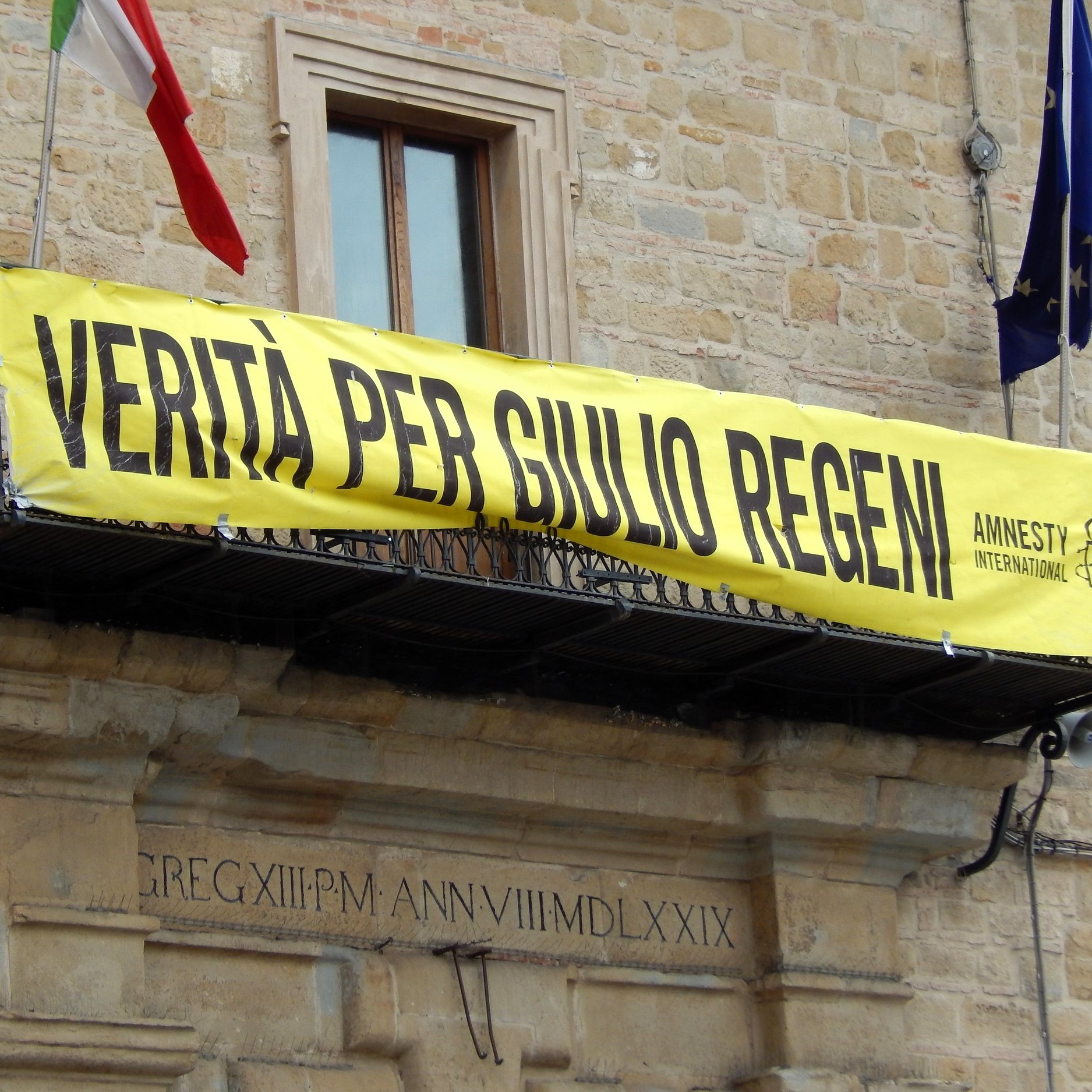 L'immagine mostra un grande striscione giallo appeso a un balcone di un edificio. Sullos striscione è scritto "VERITÀ PER GIULIO REGENI", accompagnato dal logo di Amnesty International. Nel contesto, si possono notare delle bandiere, probabilmente italiane, che sventolano. L'atmosfera suggerisce una manifestazione di sostegno e richiesta di giustizia per Giulio Regeni, un caso di rilevanza pubblica.