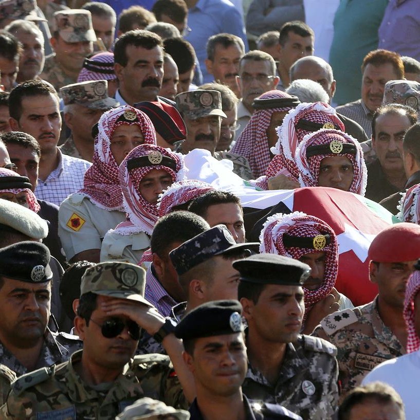 L'image montre une grande foule rassemblée. Au premier plan, des hommes portant des keffiehs (un type de couvre-chef traditionnel) entourent un cercueil recouvert d'un drapeau. Les participants semblent montrer leurs respects, et on peut également voir des militaires en uniforme dans la foule. L'atmosphère semble solennelle, indiquer une cérémonie ou des funérailles d'un individu important.