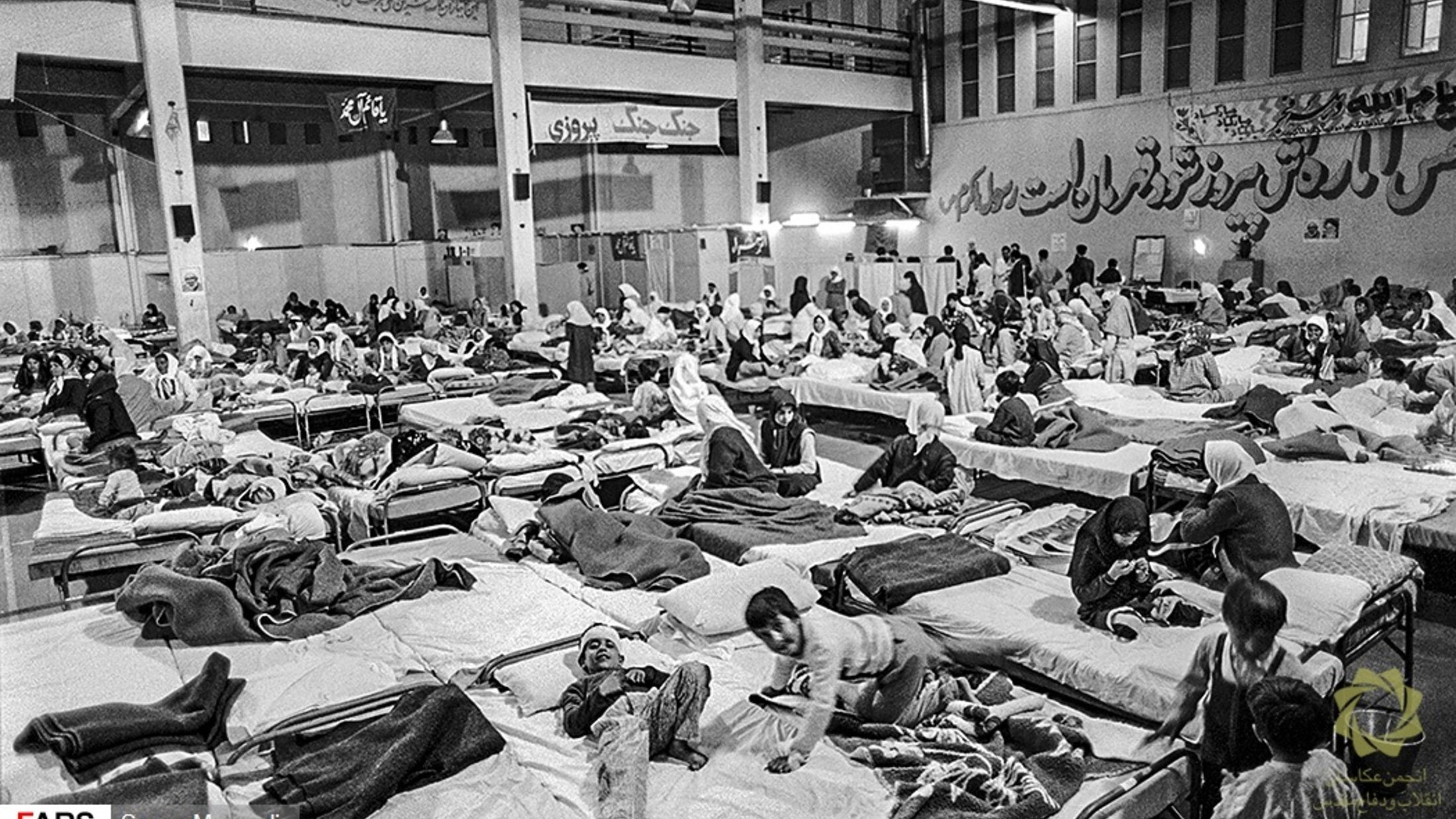 L'image montre un grand espace intérieur aménagé en salle de soins, avec de nombreux lits occupés par des personnes. On peut voir des patients allongés sur des matelas, certains semblant recevoir des soins médicaux. L'atmosphère est chargée et il y a un sentiment d'urgence, avec une présence significative de personnel soignant et de familles. Les murs portent des inscriptions en persan, ajoutant à l'authenticité du lieu.