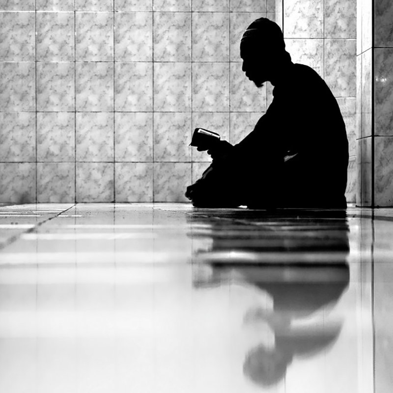 L'image montre un individu assis sur le sol, plongé dans la lecture d'un livre. La composition est en noir et blanc, créant un contraste fort entre l'ombre de la personne et l'éclat des carreaux brillants au sol. L'ambiance est calme et contemplative, et le reflet de la silhouette sur le sol ajoute une dimension introspective à la scène. Les murs en arrière-plan sont recouverts de carrelage, ce qui renforce l'aspect minimaliste de l'image.