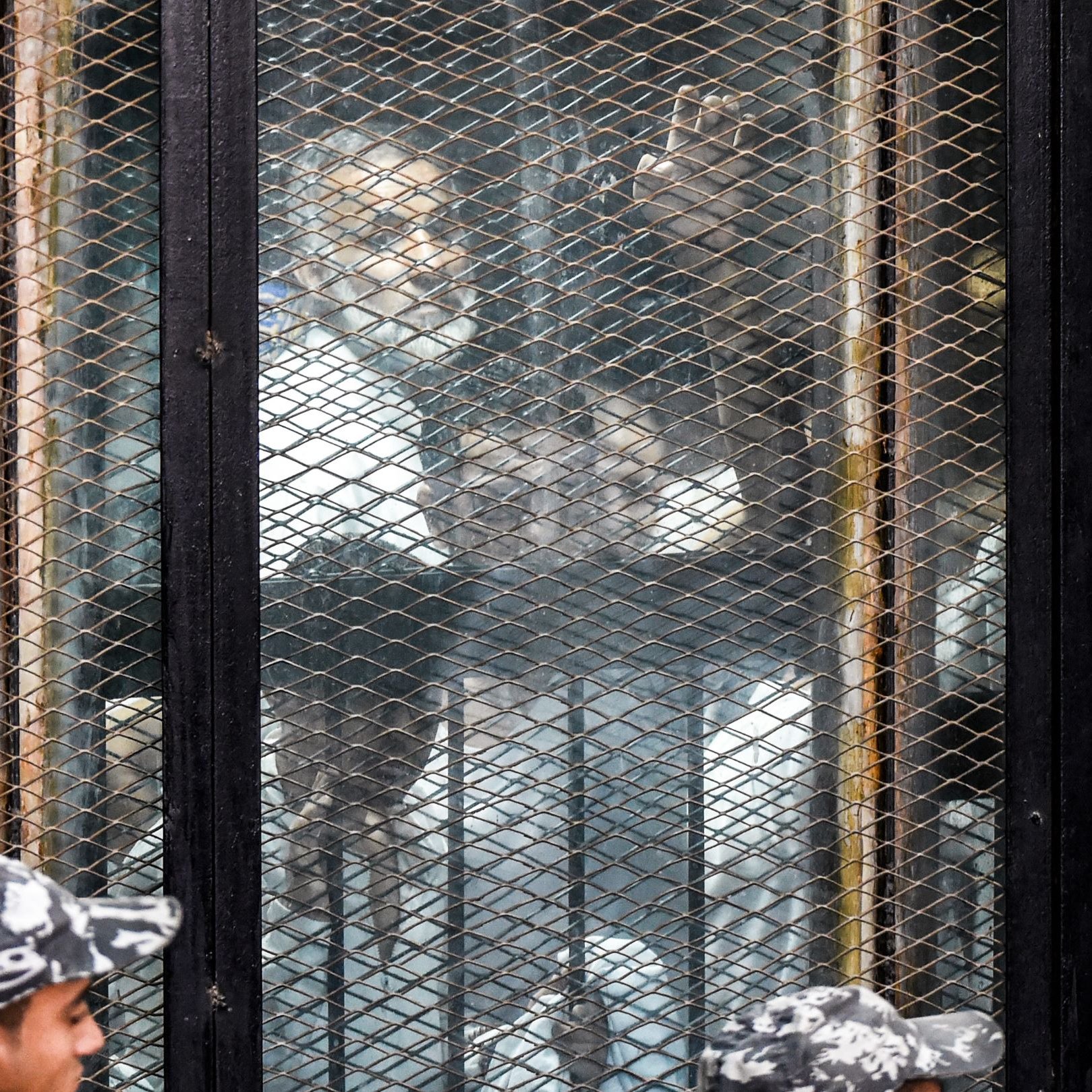 L'image montre une scène où des personnes se trouvent derrière une grille ou une clôture métallique. On aperçoit plusieurs individus, probablement en détention, qui semblent observés par des gardiens ou des forces de l'ordre en uniforme. L'atmosphère semble tendue, et il y a un mélange de visages. L'environnement donne une impression de confinement et de surveillance.