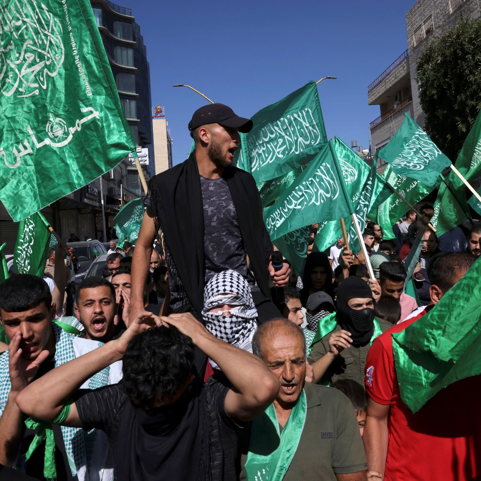 Cette image montre une manifestation animée, avec des personnes brandissant de grands drapeaux verts. On peut voir une foule de manifestants, certains sont en train de crier ou de chanter, tandis qu'un homme est porté sur les épaules de quelqu'un d'autre. L'ambiance semble énergique, et l'arrière-plan présente des bâtiments urbains.