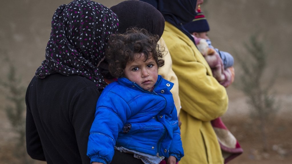 L'image montre plusieurs personnes, dont une femme tenant un enfant dans ses bras. L'enfant porte une veste bleue, et son expression semble triste ou préoccupée. Les femmes qui les entourent sont habillées de manière chaleureuse et portent des foulards. L'arrière-plan est flou, avec des éléments naturels, ce qui suggère un environnement difficile. L'ensemble de la scène évoque une atmosphère de vulnérabilité et de défi.