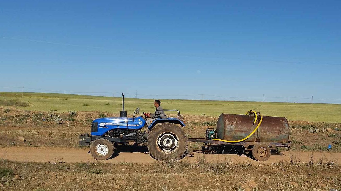 L'image montre un homme conduisant un tracteur bleu sur une route de terre. Il tire une remorque avec un grand réservoir métallique. À l'arrière-plan, on peut voir des champs verts sous un ciel dégagé. La scène évoque une ambiance rurale et agricole.