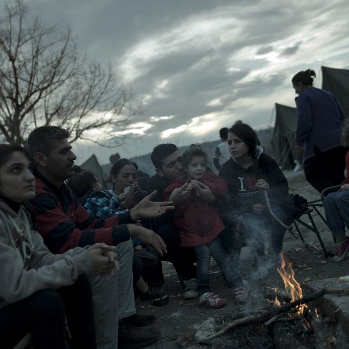 L'image montre un groupe de personnes assises autour d'un feu de camp, probablement dans un camp de réfugiés ou un site de fortune. On peut voir des adultes et des enfants, certains vêtus de manteaux pour se protéger du froid. L'ambiance semble sombre en raison des nuages menaçants dans le ciel. Les visages des personnes expriment une variété d'émotions, allant de la tristesse à l'espoir. En arrière-plan, on aperçoit des tentes qui indiquent que ce groupe vit dans des conditions temporaires.