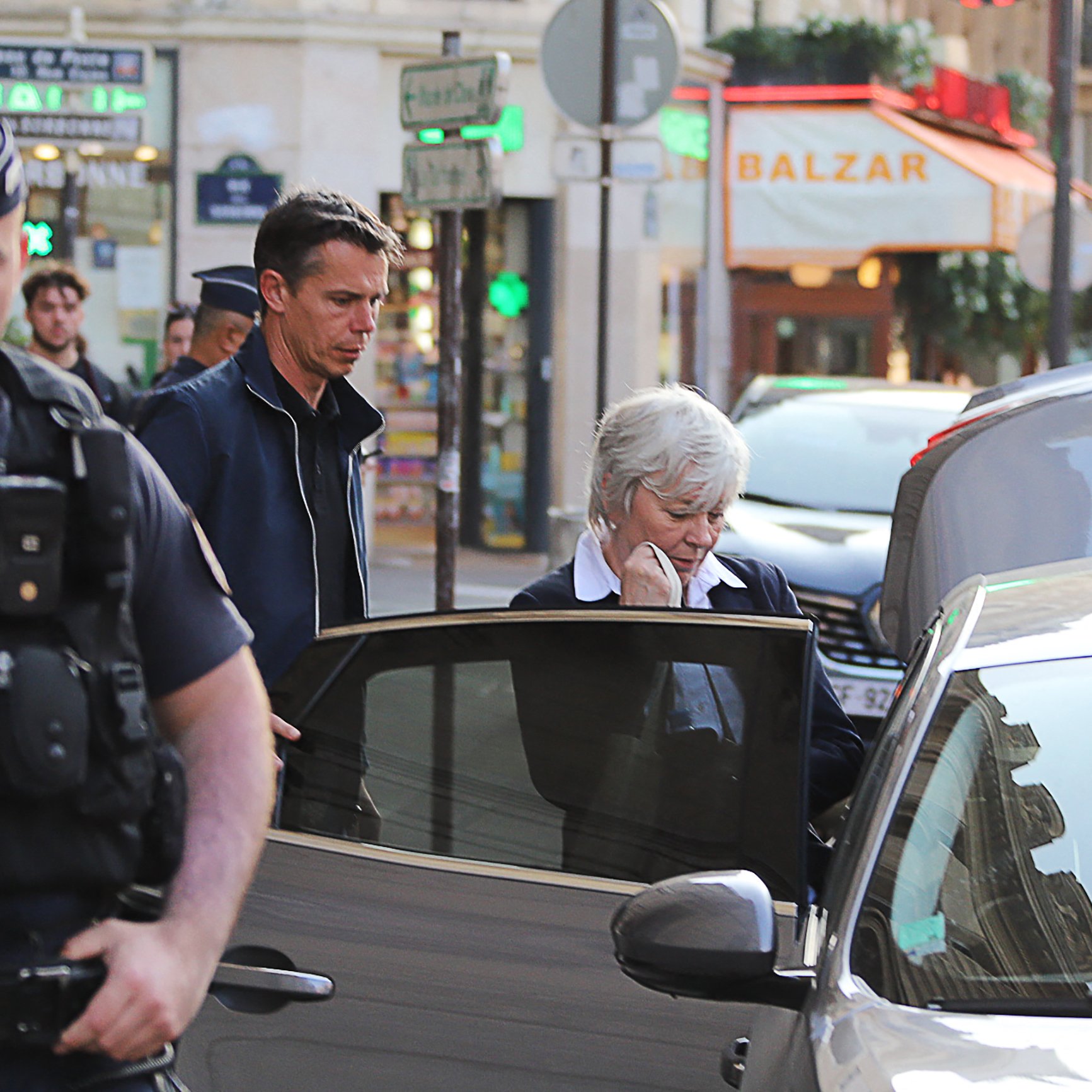 L'image montre une scène urbaine avec plusieurs personnes dans une rue. On peut voir un policier en uniforme à l'avant, surveillant la situation. À côté de lui, un homme et une femme semblent sortir d'une voiture. La femme est habillée de manière formelle et semble pensive. En arrière-plan, il y a plusieurs enseignes de magasins qui ajoutent à l'ambiance de la ville. Les lumières et l'architecture confèrent une atmosphère animée à la scène.