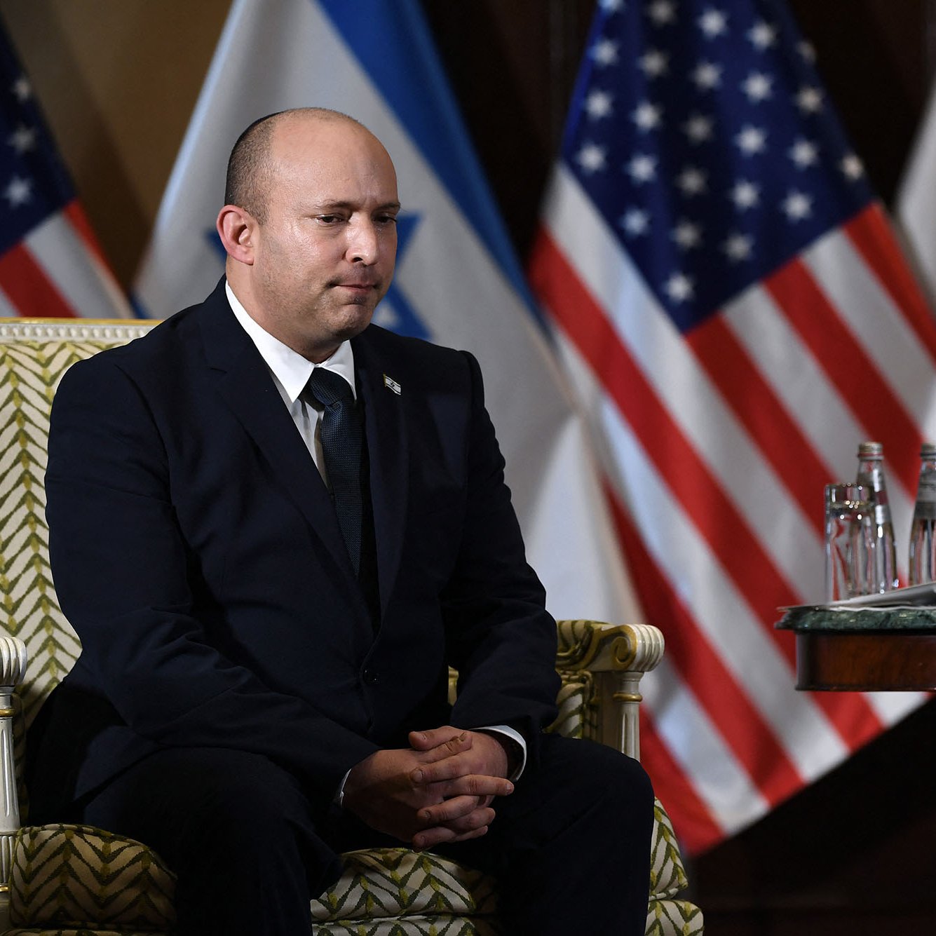 L'image montre un homme assis sur une chaise ornée, vêtu d'un costume sombre. Il a une expression sérieuse et semble concentré. À l'arrière-plan, on peut voir plusieurs drapeaux, dont ceux des États-Unis et d'Israël. Sur la table devant lui, il y a des bouteilles d'eau et des tasses. L'ambiance semble officielle et solennelle.