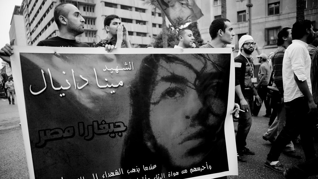 L'image montre une manifestation où des personnes portent une grande banderole. Sur la banderole, on peut voir le portrait d'une personne accompagnée d'un texte écrit en arabe, qui semble rendre hommage à cette personne. L'ambiance est chargée d'émotion, avec des manifestants en arrière-plan, et l'image est en noir et blanc, ce qui accentue la gravité de la situation. Les expressions des participants suggèrent une forte implication dans la cause défendue.