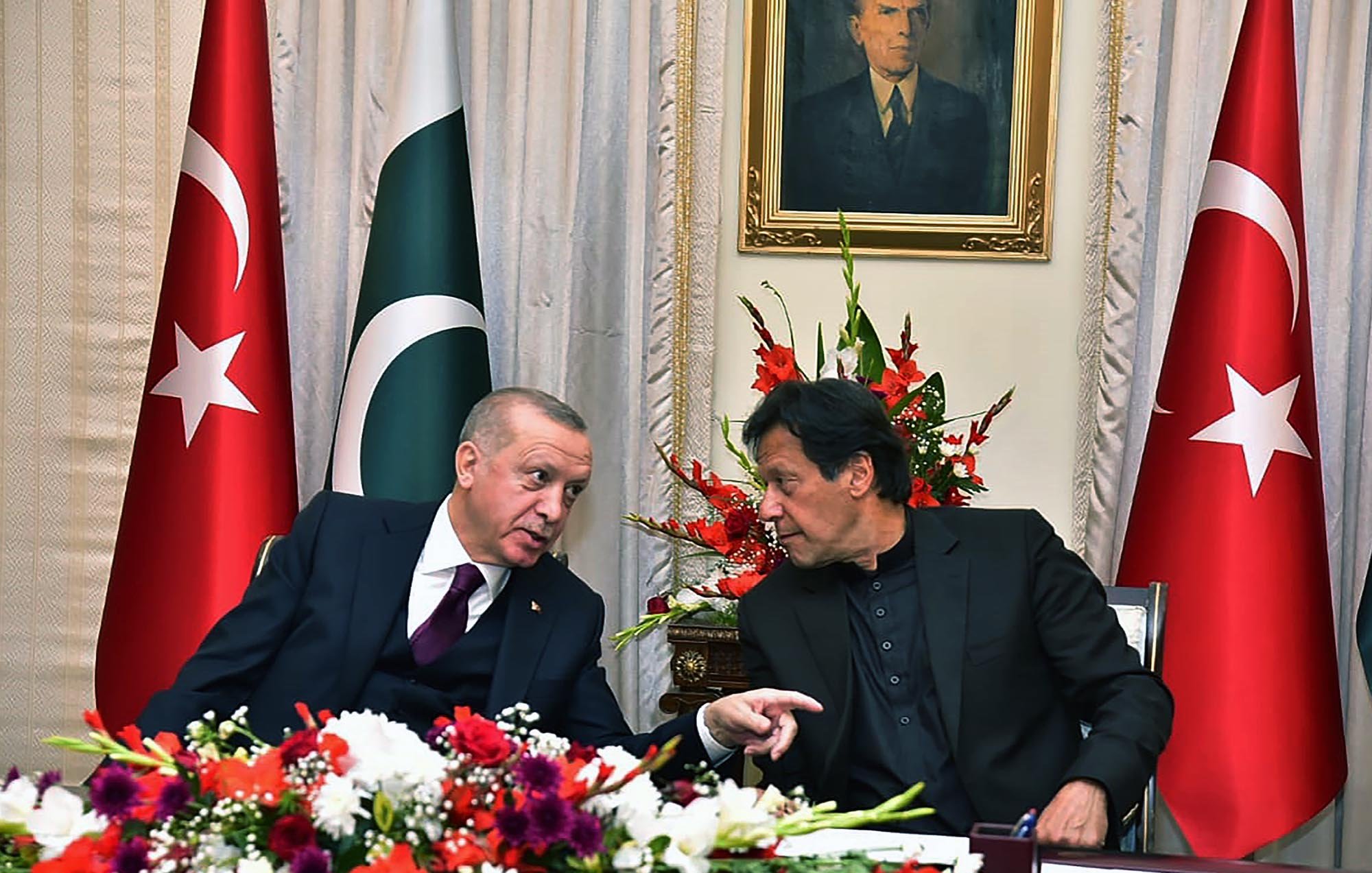 En la imagen se pueden ver a dos hombres conversando en un ambiente formal, rodeados de banderas de Turquía y Pakistán. Hay un arreglo floral colorido en la mesa, y detrás de ellos se encuentra un retrato en la pared. La escena transmite un ambiente de diplomacia y colaboración entre los dos países.