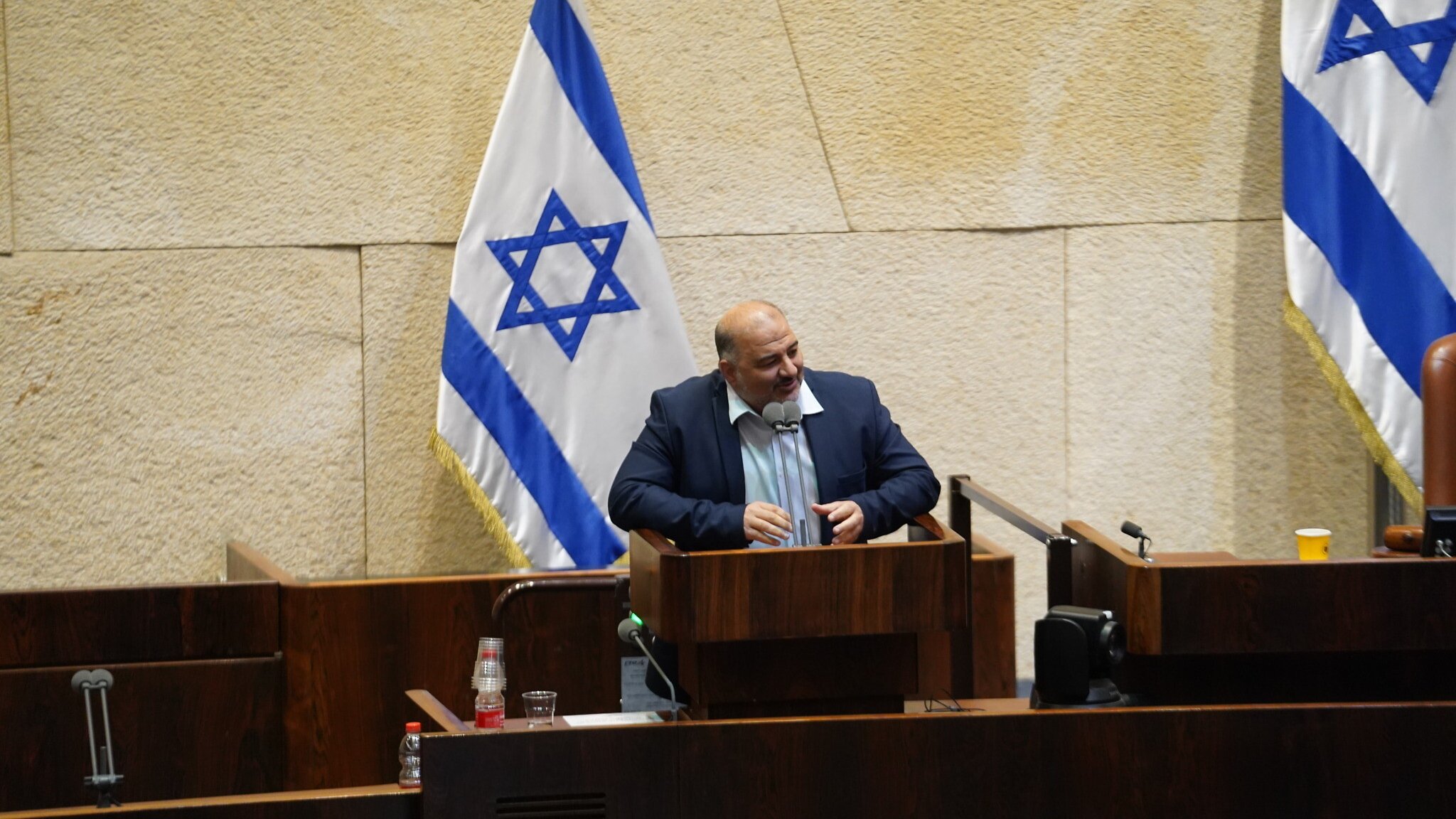 L'image montre un homme debout à un pupitre dans une salle de réunion, probablement au sein d'une assemblée ou d'un parlement, avec des drapeaux israéliens en arrière-plan. Le décor est sobre, avec des murs en pierre et du mobilier en bois. L'homme semble s'adresser à un public, exprimant peut-être son opinion sur un sujet d'actualité.