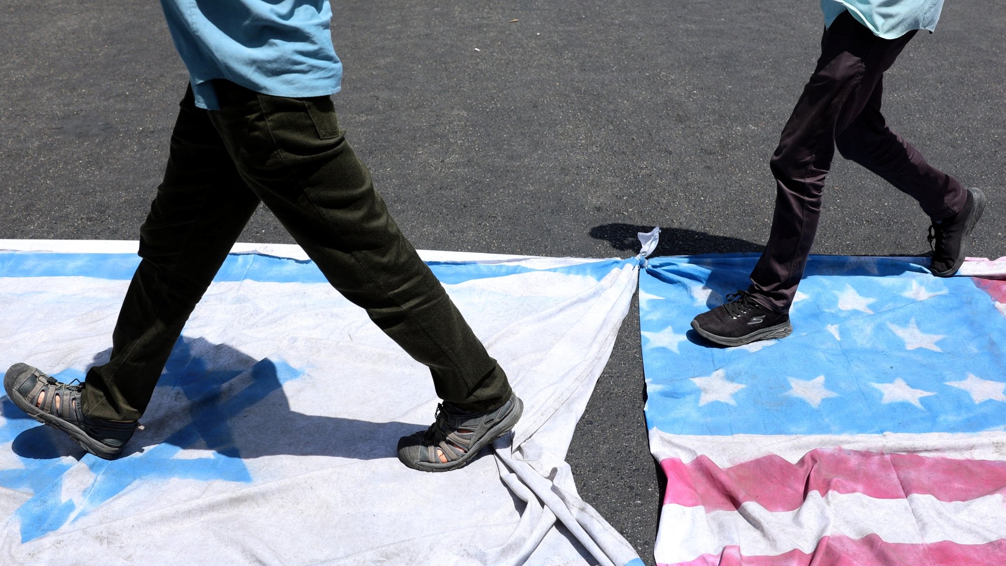 دو نفر در حال قدم زدن روی پرچم‌های دو کشور هستند. زمین زیر آن‌ها خاکی و تاریک است.