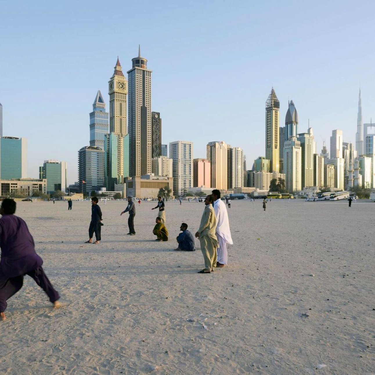 L'image montre une plage ou un espace ouvert, avec des gens qui se déplacent ou jouent dans le sable. En arrière-plan, on peut voir une ville moderne avec de grands gratte-ciel, représentant une skyline impressionnante. Les bâtiments semblent contemporains et variés en termes d'architecture. L'éclairage indique qu'il s'agit probablement d'une fin de journée, avec un ciel clair. L'atmosphère semble animée, avec des personnes profitant du lieu.