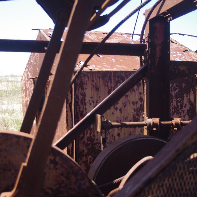 L'image montre une vue d'une structure métallique, probablement une machine ou un équipement abandonné, avec des surfaces rouillées et en décomposition. On peut voir des éléments mécaniques, tels que des poulies ou des engrenages, qui sont partiellement visibles à travers des parties de la machine. En arrière-plan, on distingue un paysage de prairies sèches, ce qui évoque une ambiance d'abandon et de dégradation. La lumière naturelle met en valeur les textures rouillées du métal.